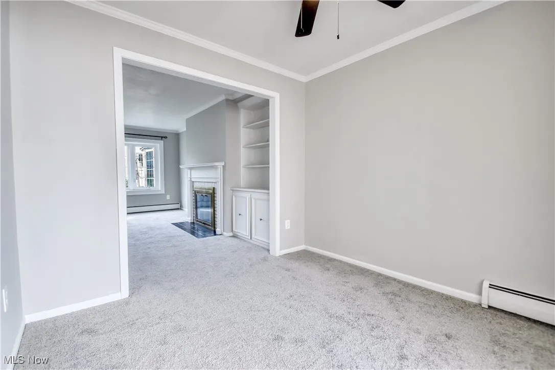 Unfurnished living room featuring built in features, ceiling fan, carpet floors, ornamental molding, and a baseboard heating unit