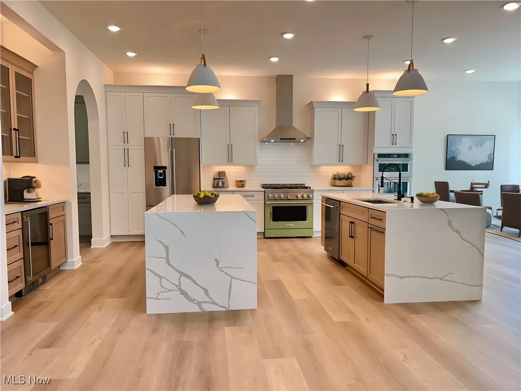 Kitchen with an island with sink, arched walkways, recessed lighting, premium appliances, and wall chimney range hood