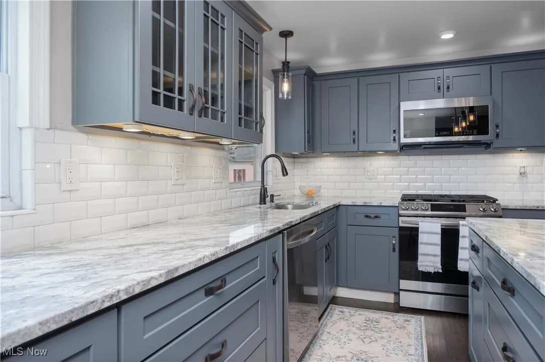 Kitchen featuring appliances with stainless steel finishes, light stone counters, glass insert cabinets, tasteful backsplash, and hanging light fixtures