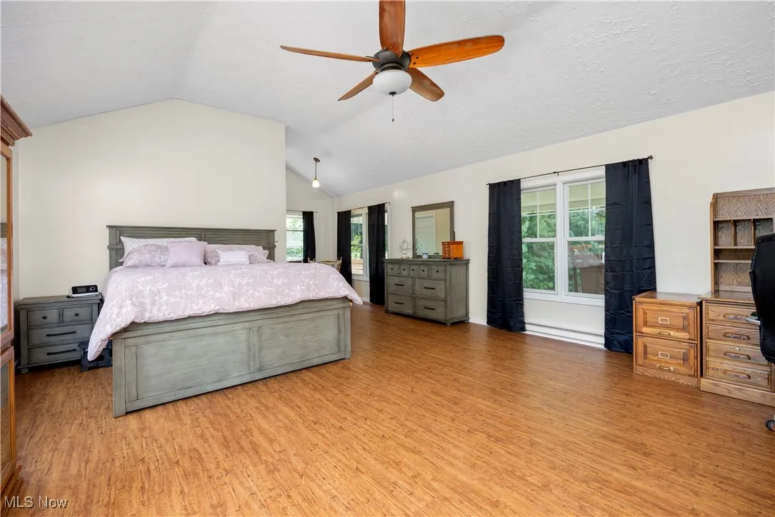 Bedroom with lofted ceiling, light wood-style floors, an office area, a baseboard heating unit, and a ceiling fan