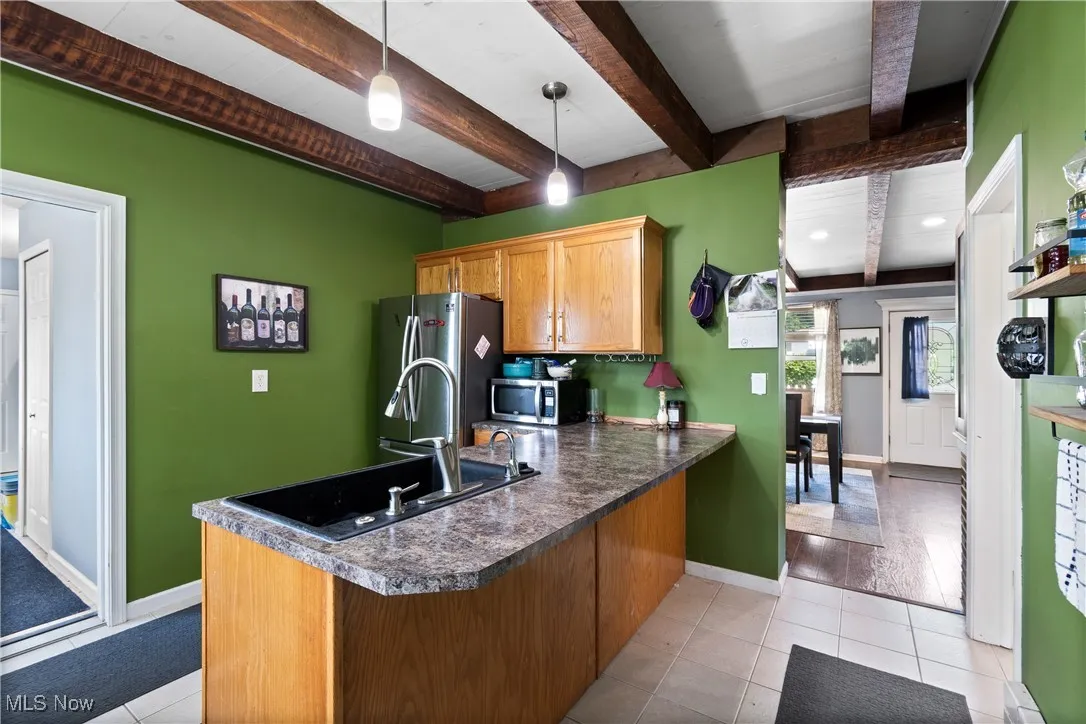 Kitchen featuring beamed ceiling, a peninsula, dark countertops, appliances with stainless steel finishes, and light tile patterned flooring