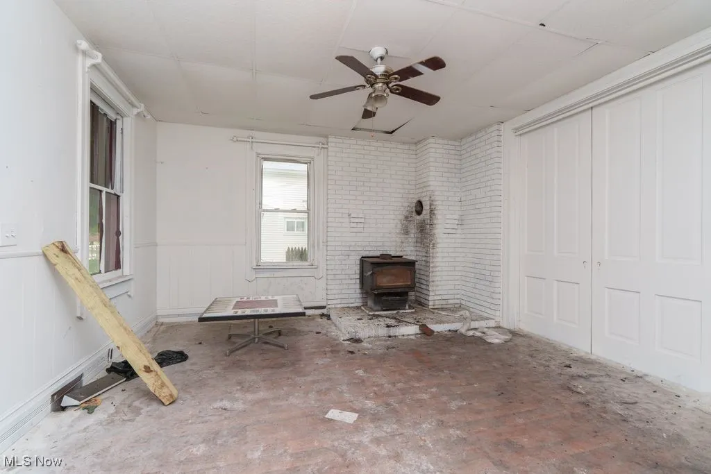 Unfurnished living room featuring a wood stove, wainscoting, a ceiling fan, and a paneled ceiling