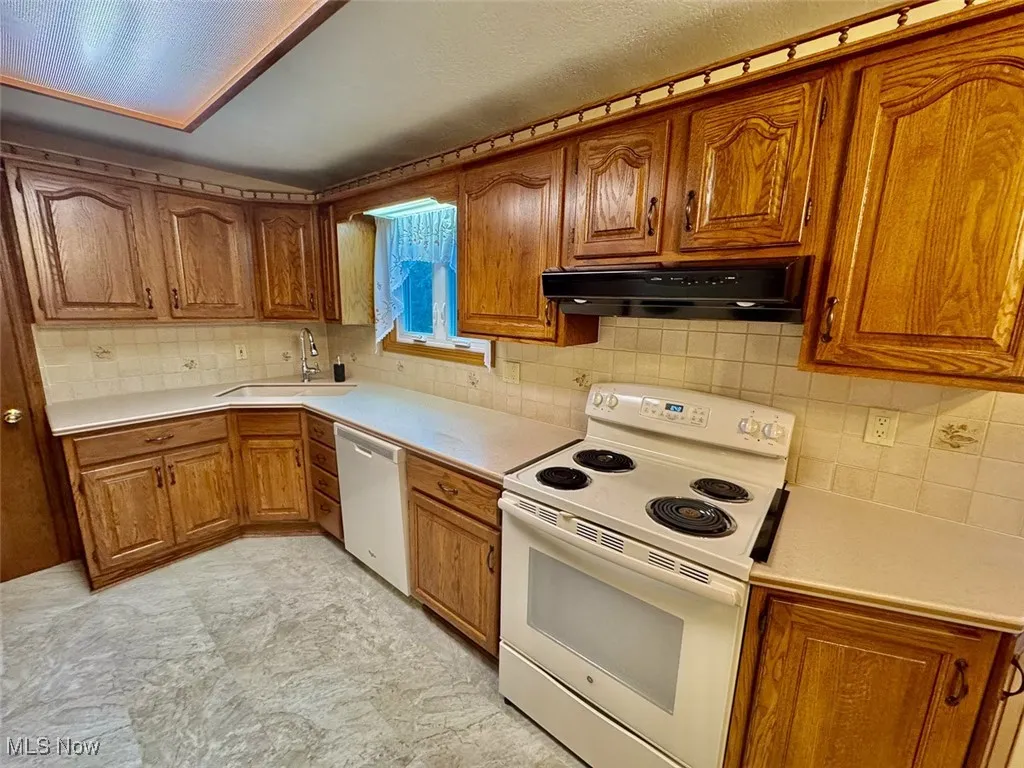 Kitchen featuring white appliances, brown cabinetry, under cabinet range hood, and tasteful backsplash