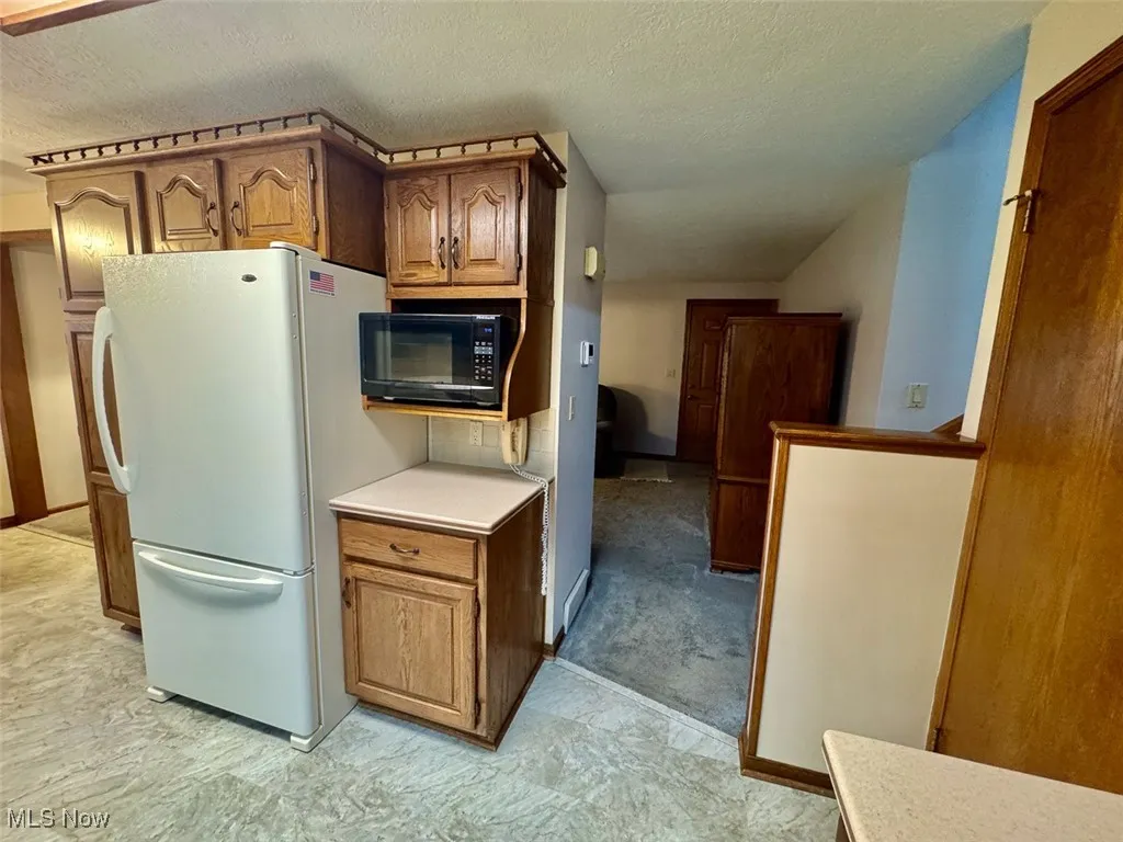 Kitchen featuring freestanding refrigerator, black microwave, light countertops, a textured ceiling, and brown cabinetry