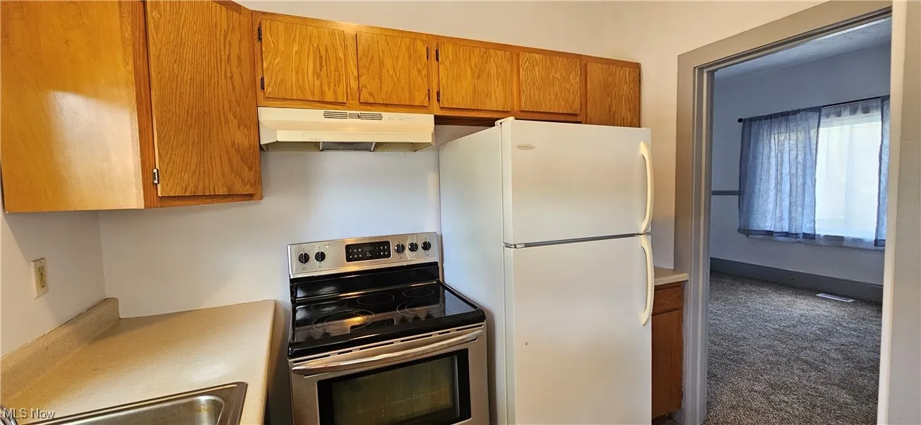 Kitchen featuring electric stove, under cabinet range hood, light countertops, and brown cabinets