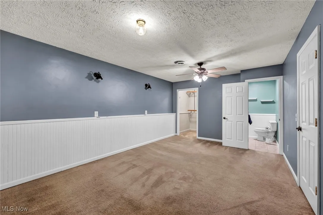 Unfurnished bedroom with a textured ceiling, wainscoting, carpet, connected bathroom, and a ceiling fan