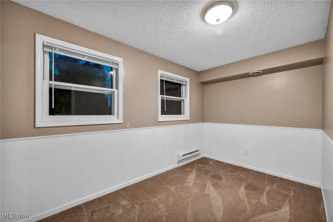 Empty room with a textured ceiling, carpet floors, wainscoting, and baseboard heating