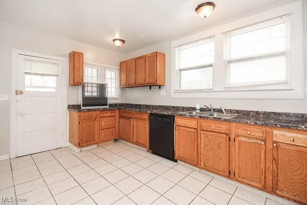 Kitchen with black dishwasher, brown cabinets, light tile patterned floors, and dark stone countertops