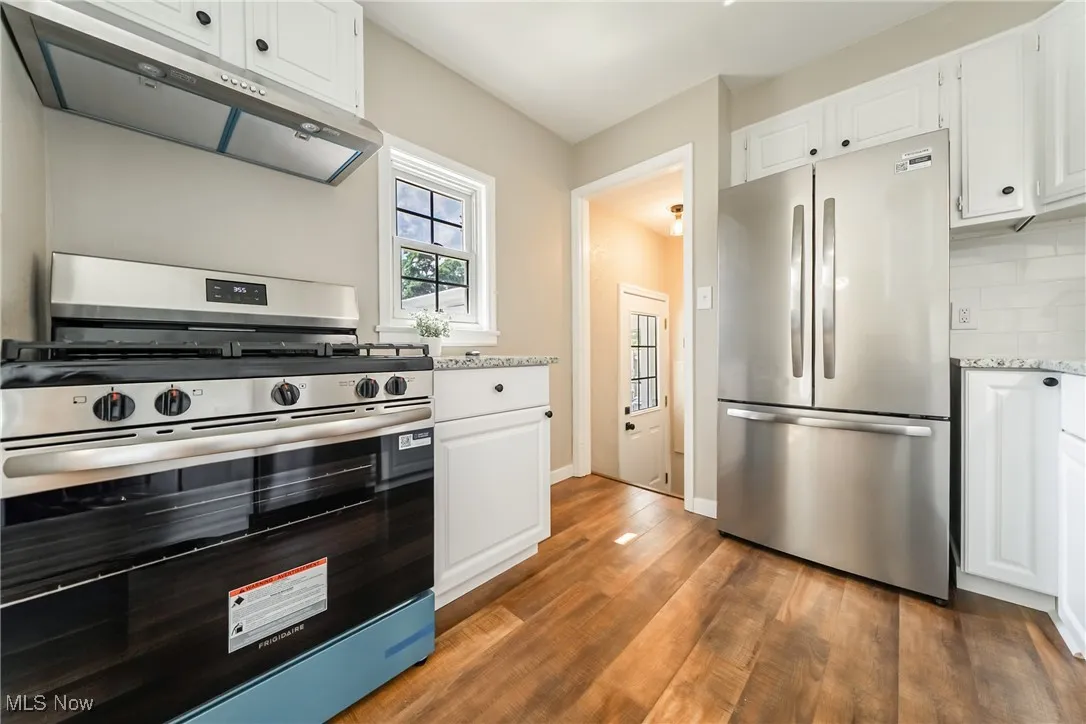 Kitchen featuring appliances with stainless steel finishes, under cabinet range hood, wood finished floors, tasteful backsplash, and white cabinets