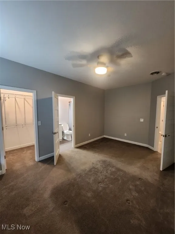 Bedroom with dark carpet, a spacious closet, ensuite bath, and a ceiling fan