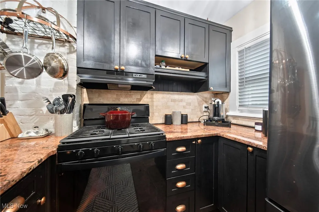 Kitchen featuring freestanding refrigerator, black range with gas stovetop, backsplash, under cabinet range hood, and light stone counters