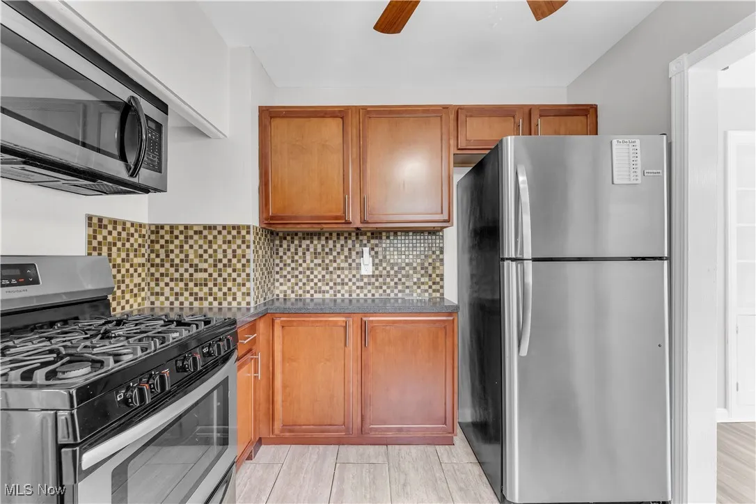 Kitchen featuring appliances with stainless steel finishes, a ceiling fan, brown cabinets, dark countertops, and tasteful backsplash