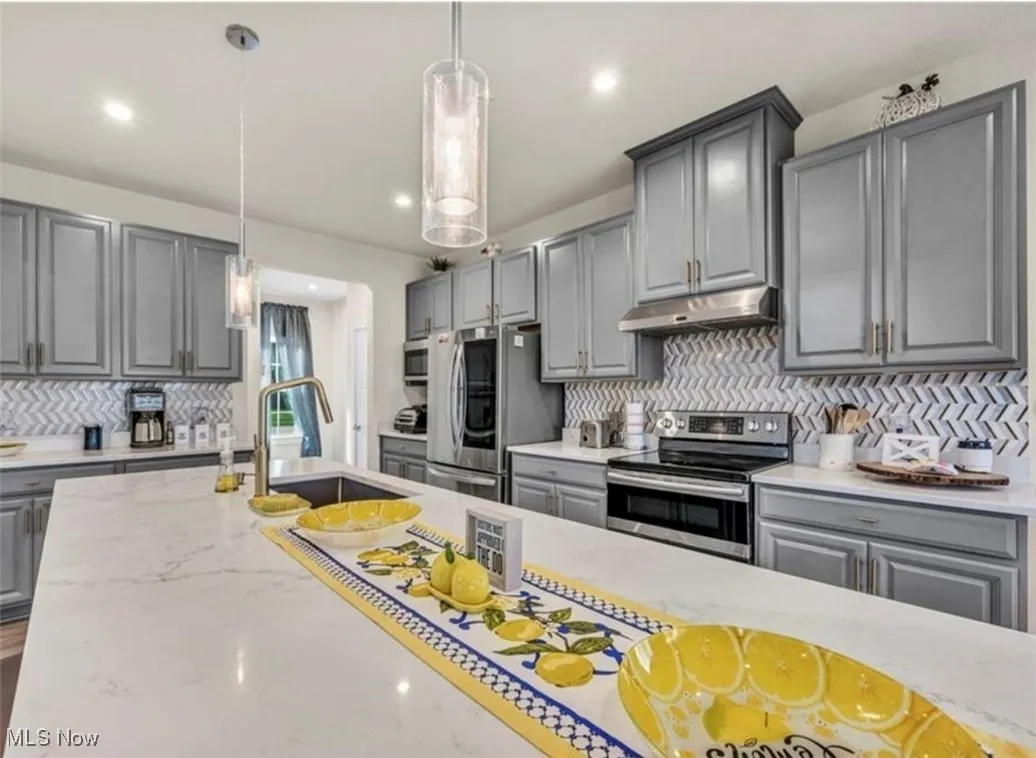Kitchen with gray cabinetry, decorative backsplash, stainless steel appliances, under cabinet range hood, and pendant lighting