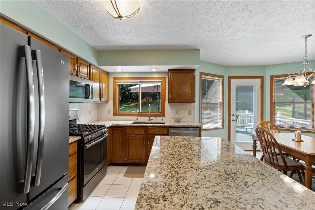Kitchen featuring stainless steel appliances, backsplash, brown cabinetry, a chandelier, and light stone countertops