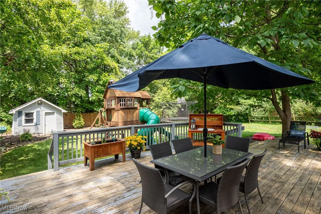 Wooden terrace featuring a playground, a fenced backyard, outdoor dining space, a storage unit, and view of wooded area