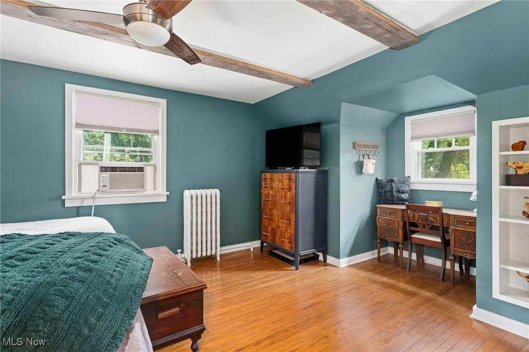 Bedroom with beam ceiling, radiator heating unit, wood finished floors, multiple windows, and ceiling fan
