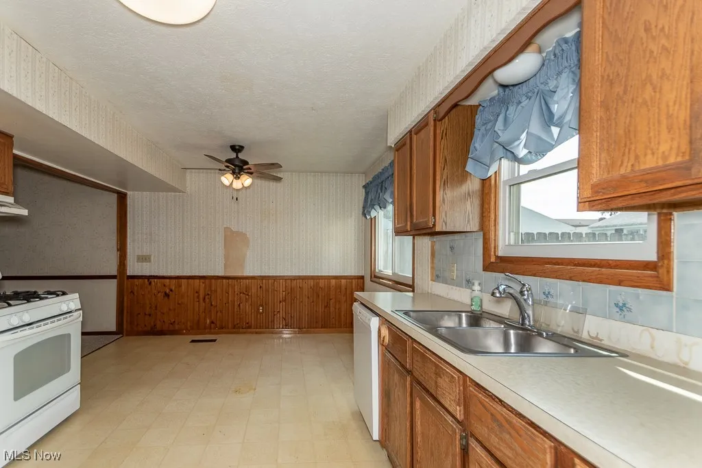 Kitchen featuring wallpapered walls, wainscoting, light flooring, white appliances, and brown cabinetry
