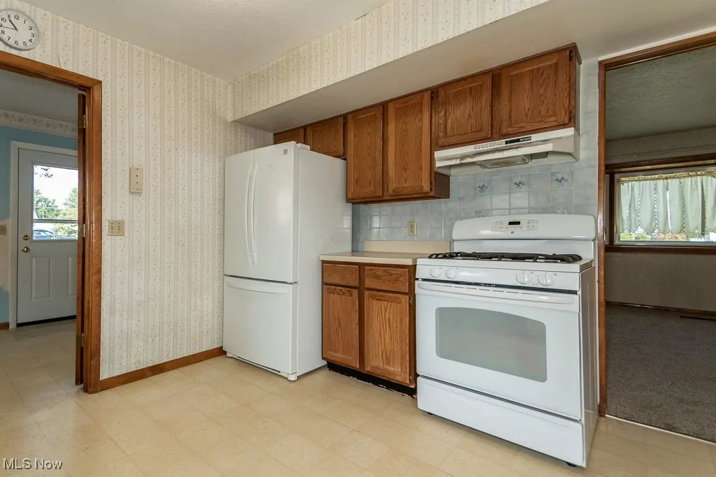 Kitchen featuring light flooring, white appliances, brown cabinets, under cabinet range hood, and wallpapered walls