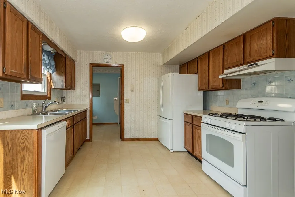 Kitchen featuring white appliances, wallpapered walls, light floors, under cabinet range hood, and brown cabinets