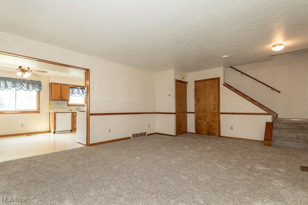 Unfurnished living room with wallpapered walls, light colored carpet, stairway, ceiling fan, and a textured ceiling