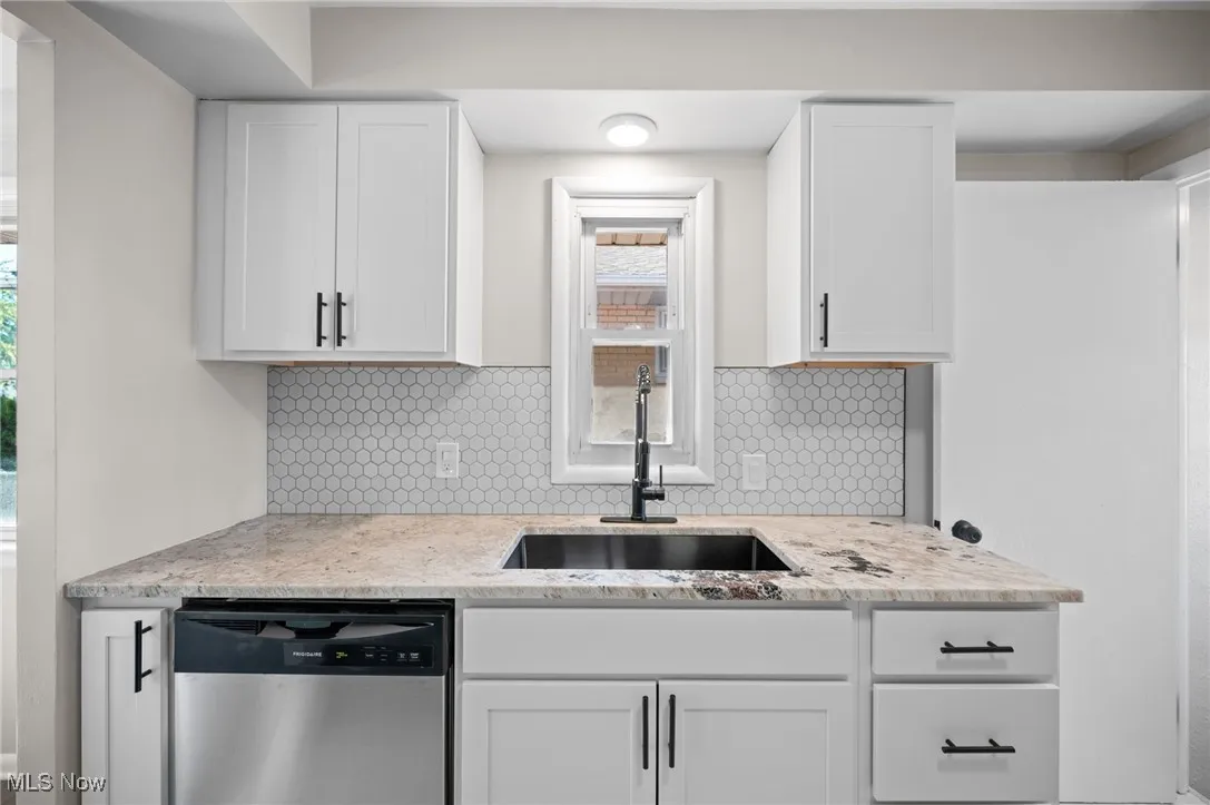 Kitchen with stainless steel dishwasher, white cabinetry, backsplash, and light stone countertops