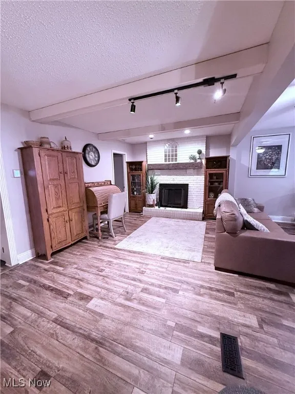 Living room with track lighting, beam ceiling, a brick fireplace, light wood-type flooring, and a textured ceiling
