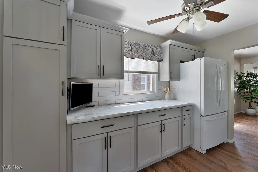 Kitchen with backsplash, wood finished floors, a ceiling fan, freestanding refrigerator, and light stone counters