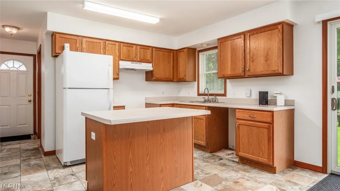 Kitchen with freestanding refrigerator, a center island, brown cabinetry, under cabinet range hood, and light countertops