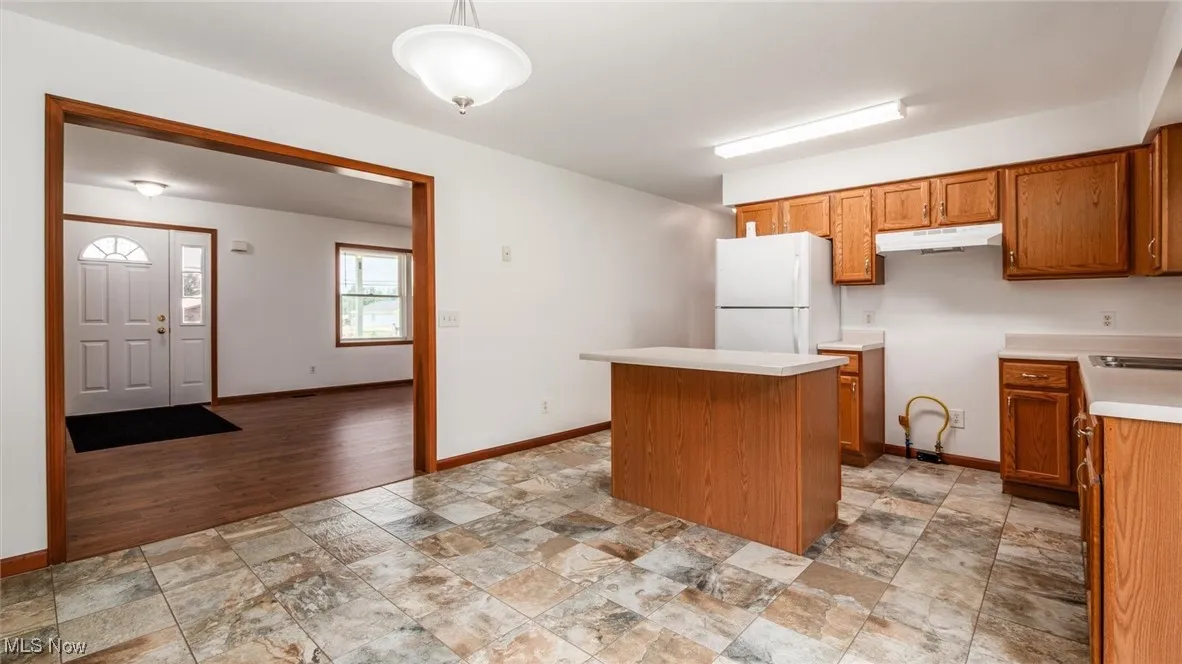 Kitchen featuring freestanding refrigerator, a center island, brown cabinets, stone finish flooring, and light countertops