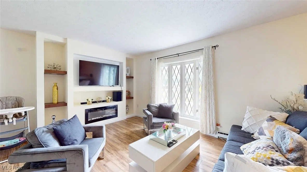 Living room featuring a glass covered fireplace, built in shelves, light wood finished floors, a textured ceiling, and a baseboard radiator