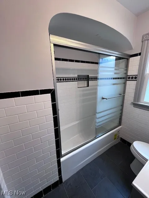 Bathroom with tile walls, combined bath / shower with glass door, tile patterned flooring, and wainscoting