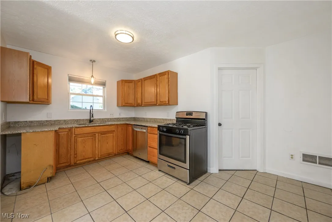 Kitchen with stainless steel appliances, brown cabinetry, a textured ceiling, light tile patterned flooring, and decorative light fixtures