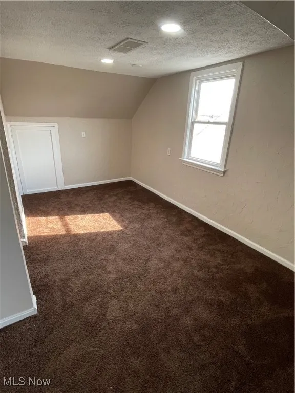 Additional living space with a textured ceiling, dark colored carpet, lofted ceiling, and a textured wall