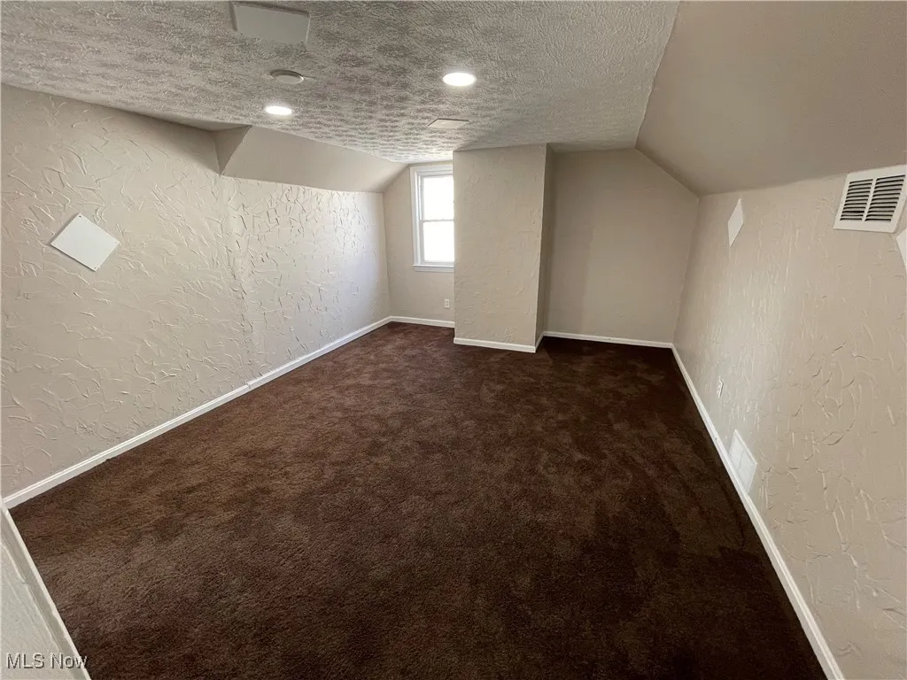 Additional living space featuring a textured wall, a textured ceiling, dark colored carpet, and lofted ceiling