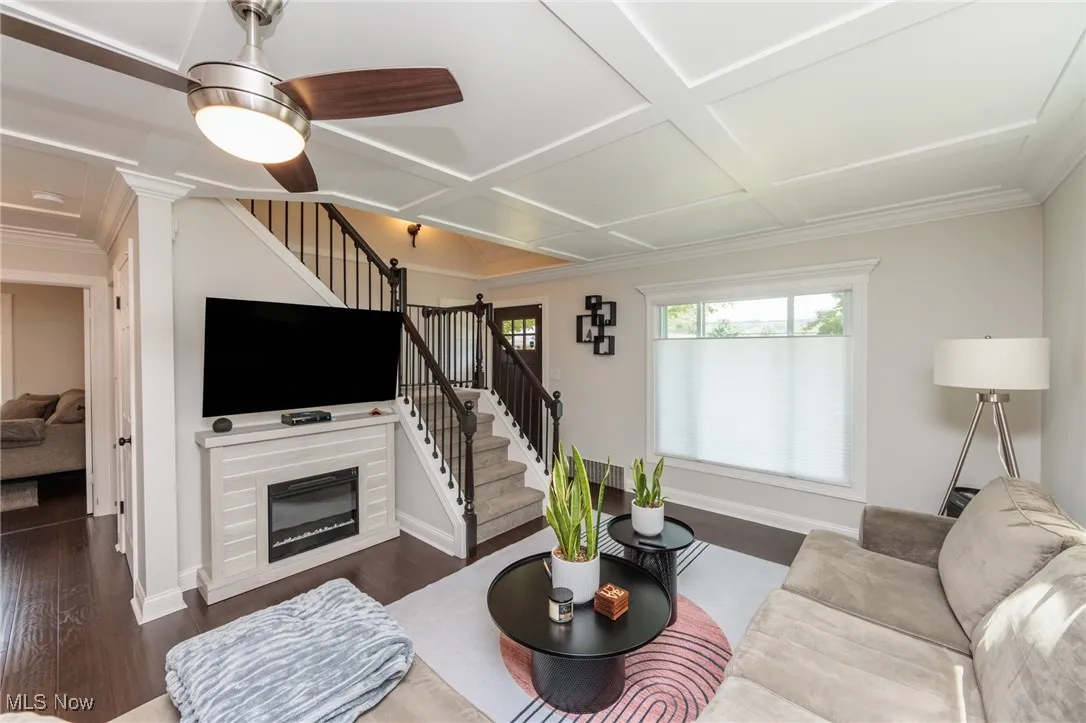 Living room featuring stairway, dark wood-style flooring, coffered ceiling, a glass covered fireplace, and crown molding