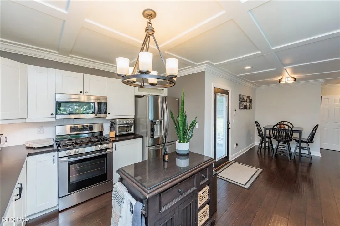 Kitchen with dark countertops, appliances with stainless steel finishes, a chandelier, dark wood-type flooring, and coffered ceiling