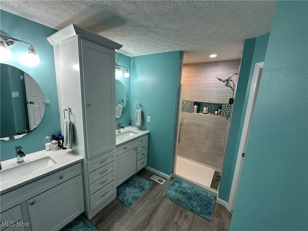 Full bathroom with a textured ceiling, a tile shower, wood finished floors, and two vanities