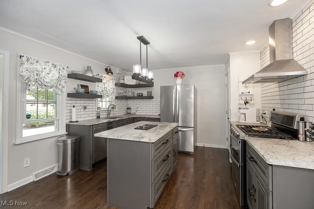 Kitchen with gray cabinetry, open shelves, stainless steel appliances, wall chimney exhaust hood, and light stone counters