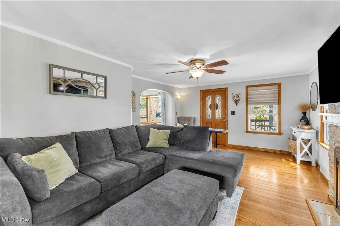 Living room with arched walkways, light wood-type flooring, crown molding, a ceiling fan, and a fireplace