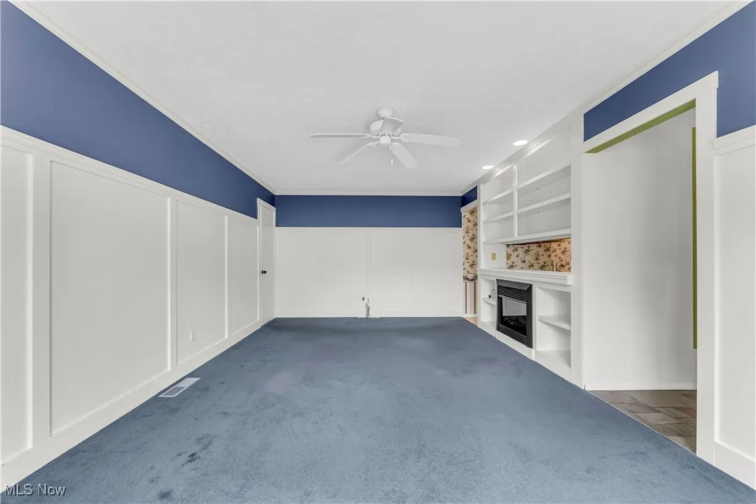Unfurnished living room featuring a decorative wall, a ceiling fan, a fireplace, a wainscoted wall, and crown molding