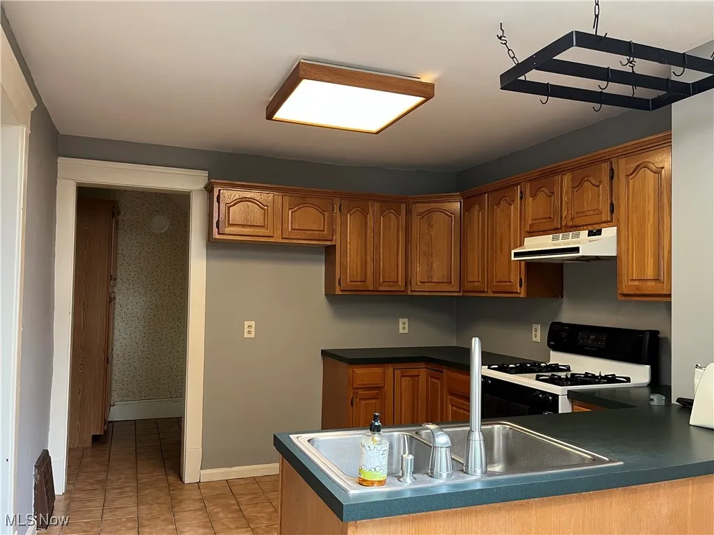 Kitchen with white gas stove, a peninsula, brown cabinetry, under cabinet range hood, and wallpapered walls