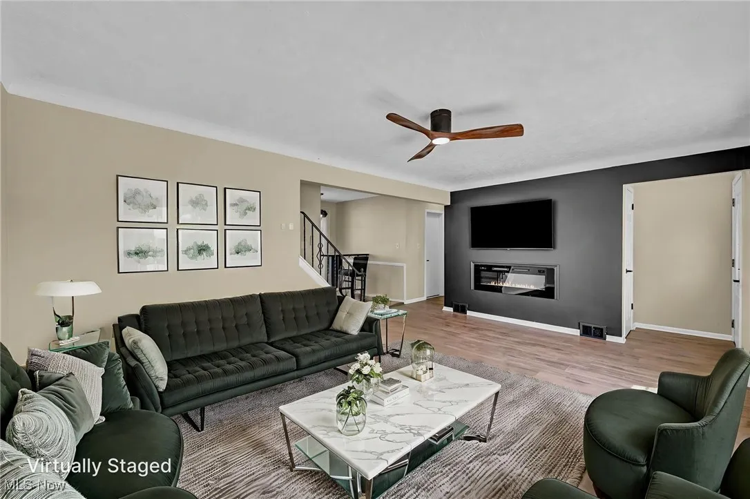 Living area with a glass covered fireplace, wood finished floors, a ceiling fan, and stairs