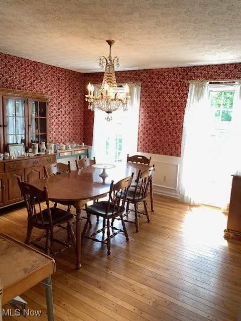 Dining area with light wood finished floors, wallpapered walls, a chandelier, a wainscoted wall, and a textured ceiling