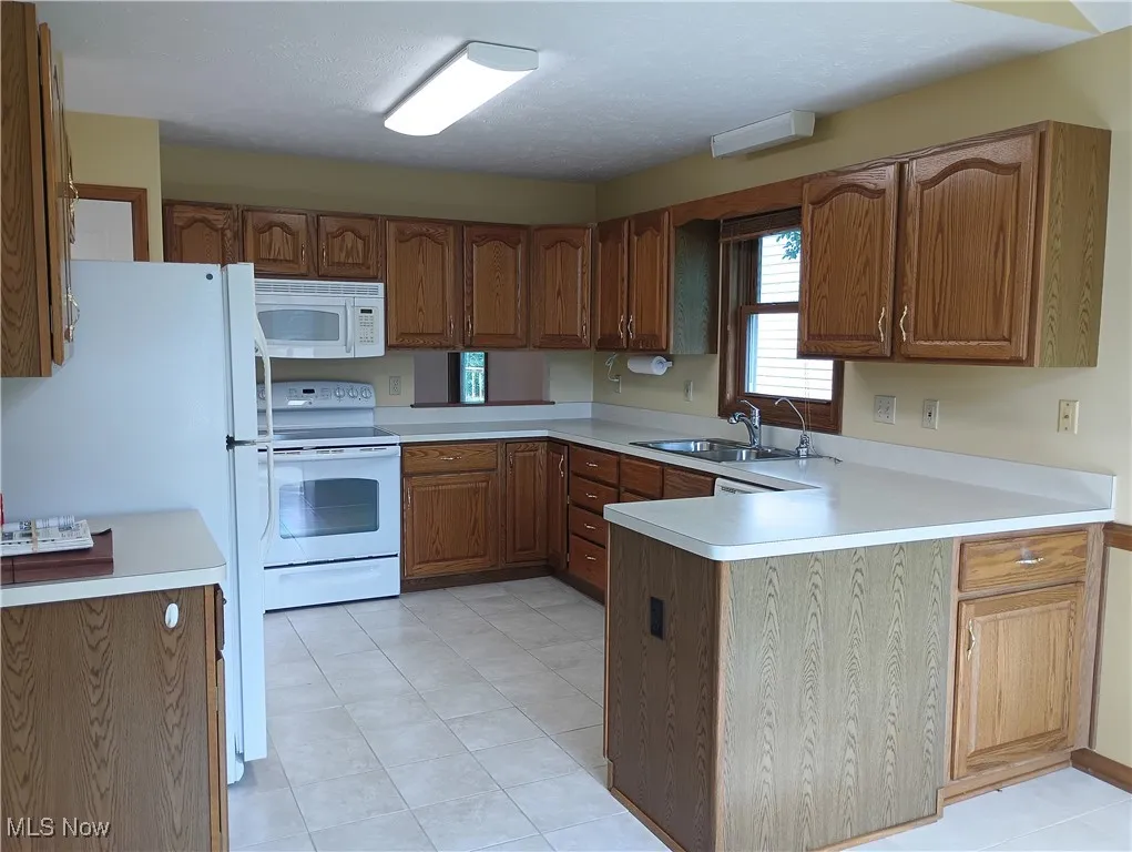 Kitchen with white appliances, a peninsula, light countertops, brown cabinetry, and light tile patterned floors