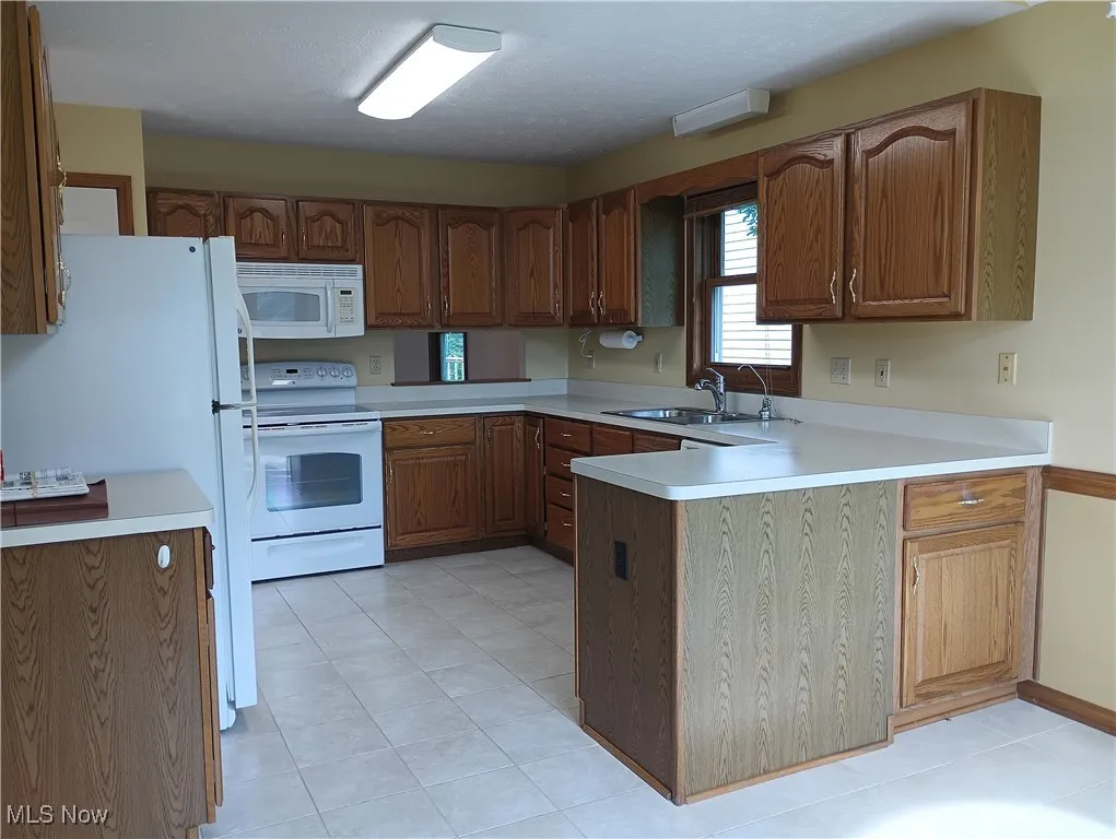 Kitchen with white appliances, a peninsula, light countertops, light tile patterned floors, and brown cabinets