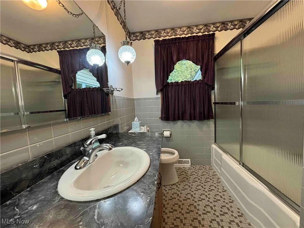 Full bath with tile walls, vanity, bath / shower combo with glass door, and wainscoting