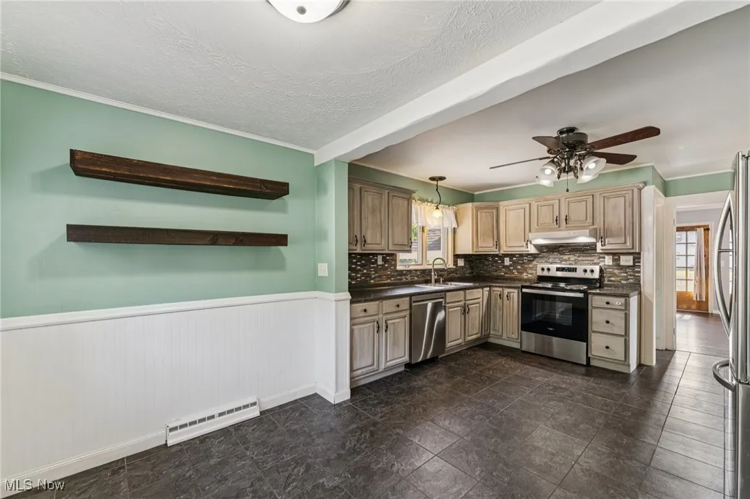 Kitchen with dark countertops, wainscoting, appliances with stainless steel finishes, a ceiling fan, and under cabinet range hood