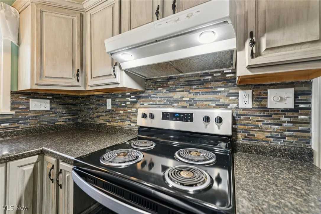 Kitchen featuring under cabinet range hood, stainless steel electric stove, backsplash, dark countertops, and light brown cabinetry
