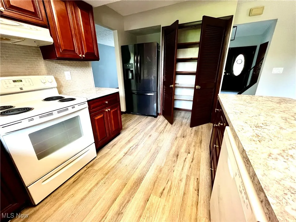 Kitchen featuring white electric range oven, reddish brown cabinets, black fridge with ice dispenser, light wood finished floors, and under cabinet range hood
