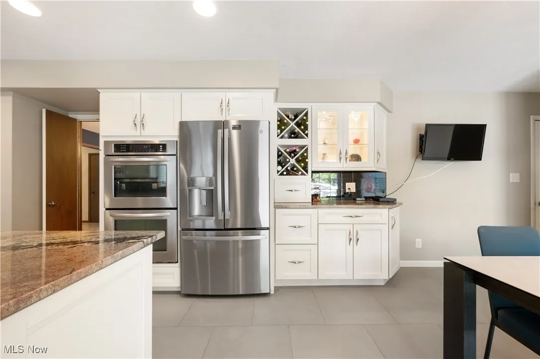 Kitchen with appliances with stainless steel finishes, stone countertops, white cabinetry, light tile patterned flooring, and glass insert cabinets